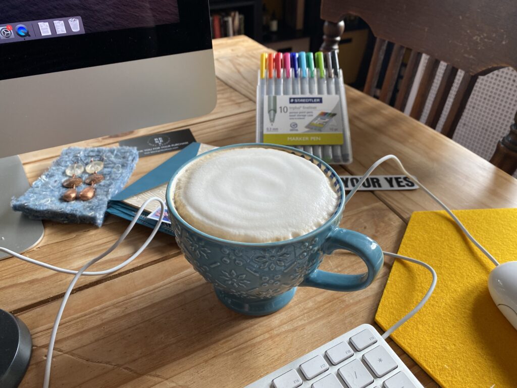 A blue coffee cup sits on a wooden table, full of latte. You can see pens, and the edge of a computer screen and keyboard.
