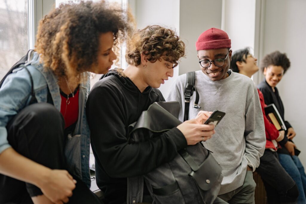 Three students look at a phone, held by the student in the middle of the photo. The student on the right is smiling