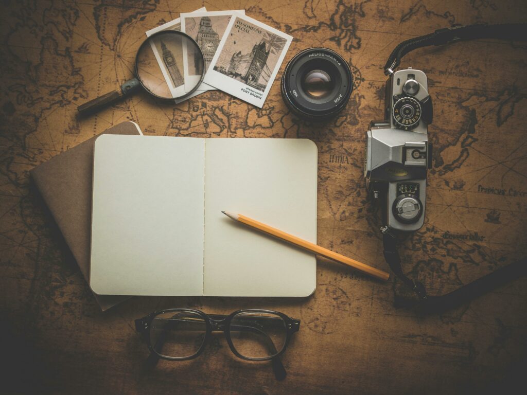 A photograph showing an open notebook, a pencil, a magnifying glass, a camera, and three polaroid pictures sitting on top of an old, sepia-toned map